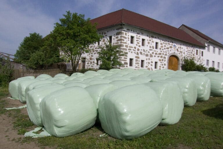 Bild von mehreren Silageballen, die vor einer weißen Feldstein-Scheune mit rotem Dach platziert sind.