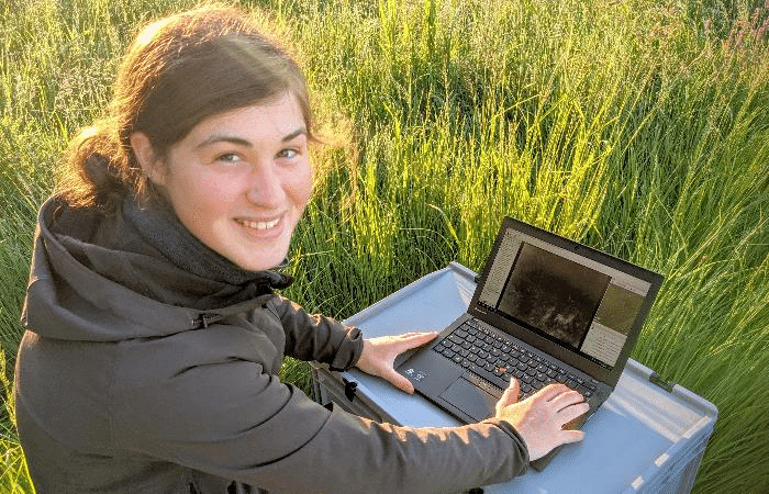 Bild von einer Person, die im Feld an einem Laptop arbeitet, auf dem die Anwendung POIStudio geöffnet ist. Die Person sieh direkt in die Kamera und lächelt.